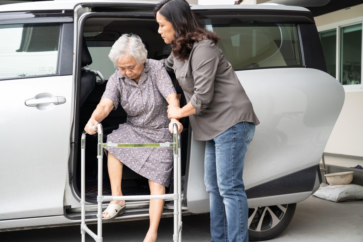 Family caregiver helping an elderly woman with a walker lower herself onto the seat of a minivan