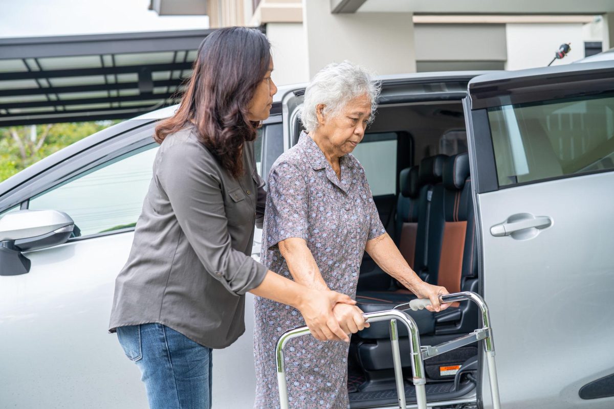 Caregiver guiding an elderly woman as she steps out of a minivan and steadies herself on a walker
