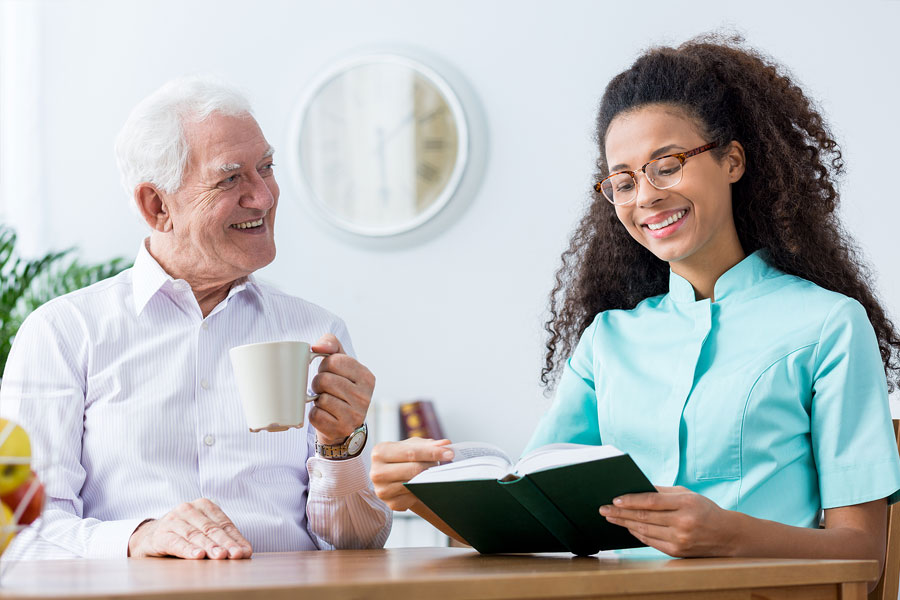 Caregiver reading book to elderly man while he drinks a cup of coffee.