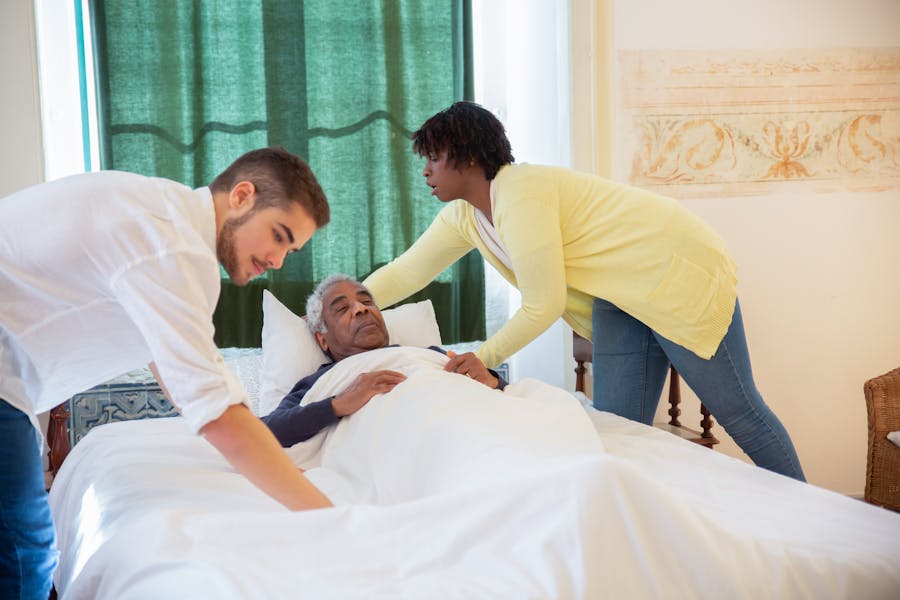 Two caregivers helping an elderly man get in bed